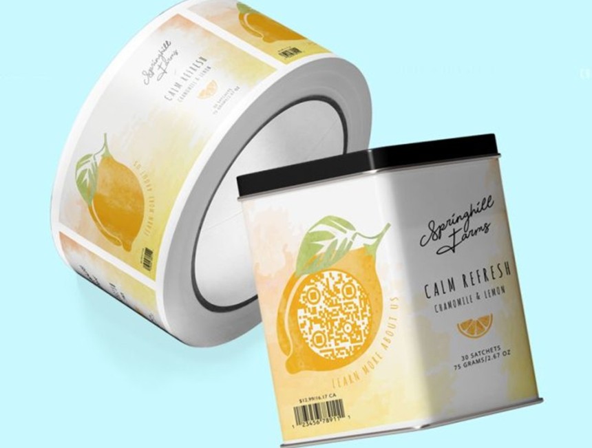 Opakowanie herbaty „Springhill Farms – Calm Refresh Chamomile & Lemon” z dużą ilustracją cytryny, w której środku znajduje się kod QR z napisem wzdłuż krawędzi „Learn more about us”; w tle rolka etykiet z tym samym projektem.
