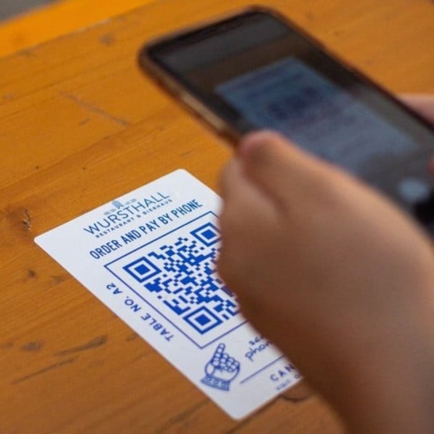 Zbliżenie dłoni trzymającej smartfon nad kartą z niebieskim kodem QR na drewnianym stoliku; na karcie widoczny jest napis po angielsku zachęcający do zamawiania i płacenia telefonem w lokalu gastronomicznym.
