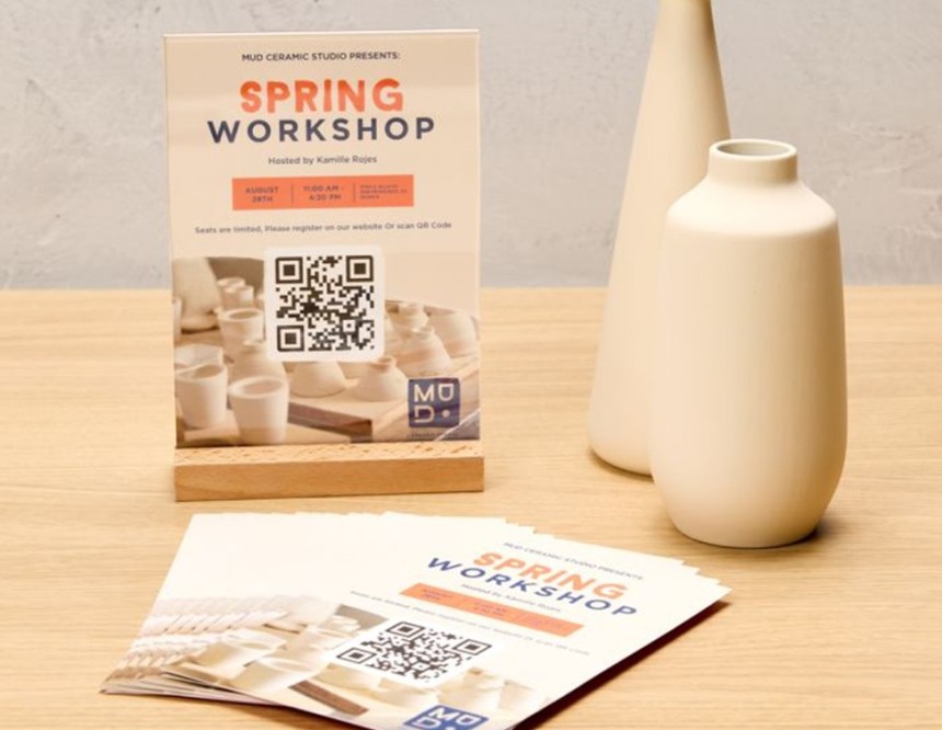 Stojąca na blacie informacja o warsztatach ceramiki „Spring Workshop” z datą, godziną i miejscem, na której centralnie umieszczono kod QR do rejestracji; obok leżą ulotki z tym samym projektem i stoją dwie jasne wazony.