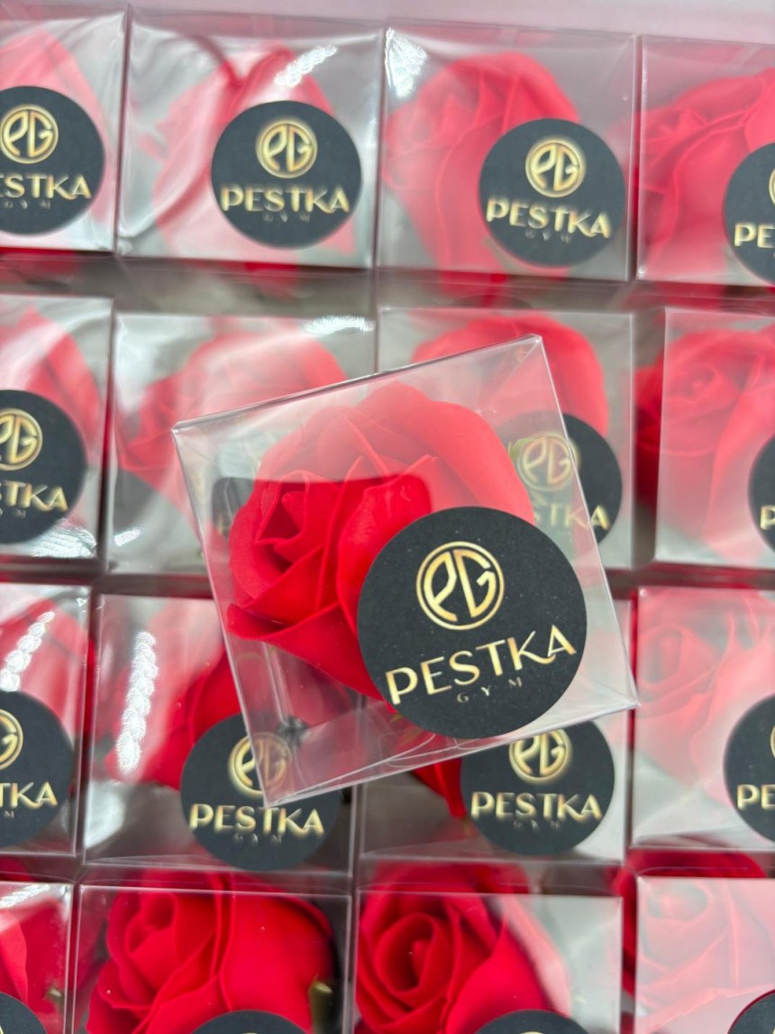  Upominek firmowy: czerwona, mydlana róża w przezroczystym pudełeczku. Na pudełku naklejono czarną, okrągłą naklejkę ze złotym logo i napisem: "PESTKA GYM". W tle widać więcej takich samych upominków.