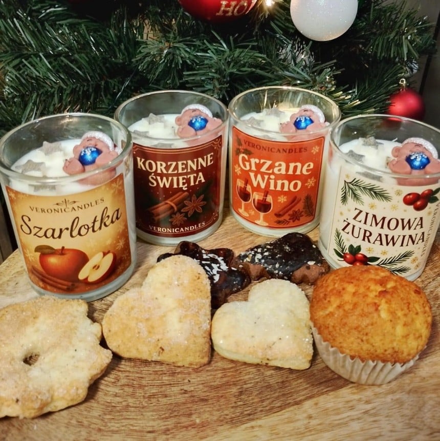 Świąteczna kompozycja z czterema świecami VeroniCandles. Każda świeca ma etykietę z nazwą zapachu (Szarlotka, Korzenne Święta, Grzane Wino, Zimowa Żurawina) i jest ozdobiona woskową figurką misia. Obok świec leżą domowe ciastka, a w tle widać oświetloną choinkę.