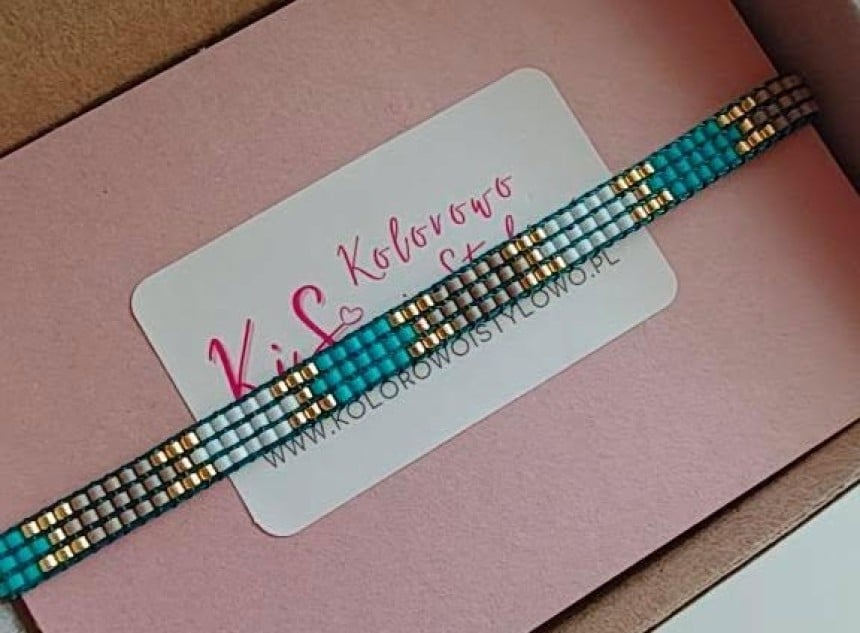 Ręcznie robiona bransoletka z drobnych, turkusowych, srebrnych i złotych koralików. Leży w pudełku na różowej podkładce z logo "KiS Kolorowo i Stylowo" i adresem strony. W rogu widoczny jest palec z różowym manicurem.