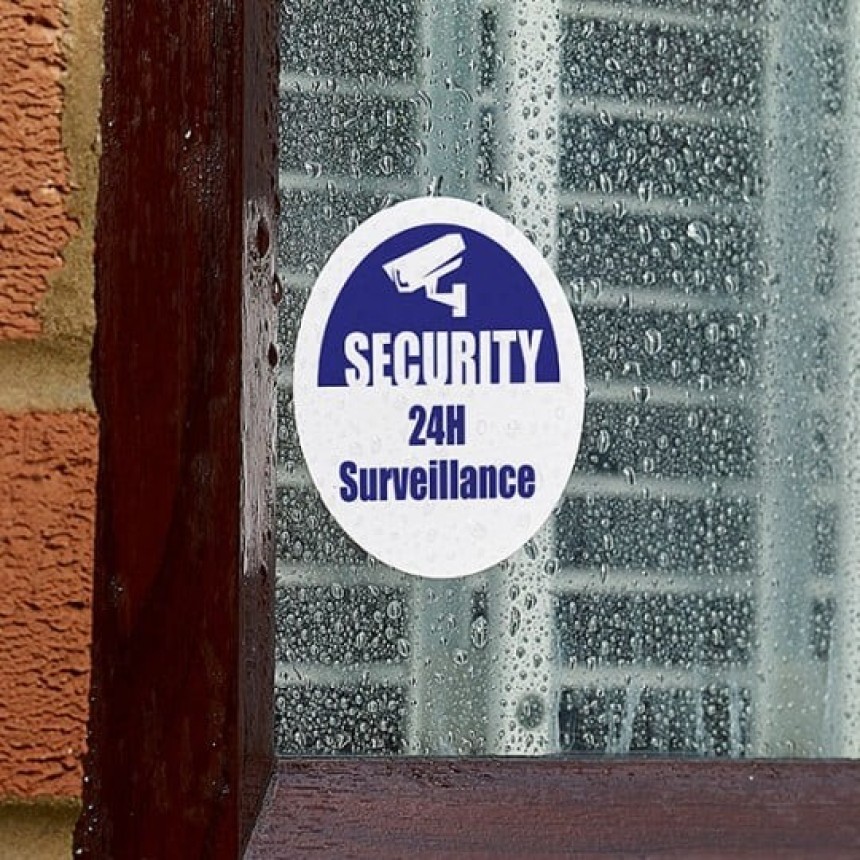 Naklejka na oknie z ikoną kamery i napisem: „SECURITY 24H Surveillance”.
