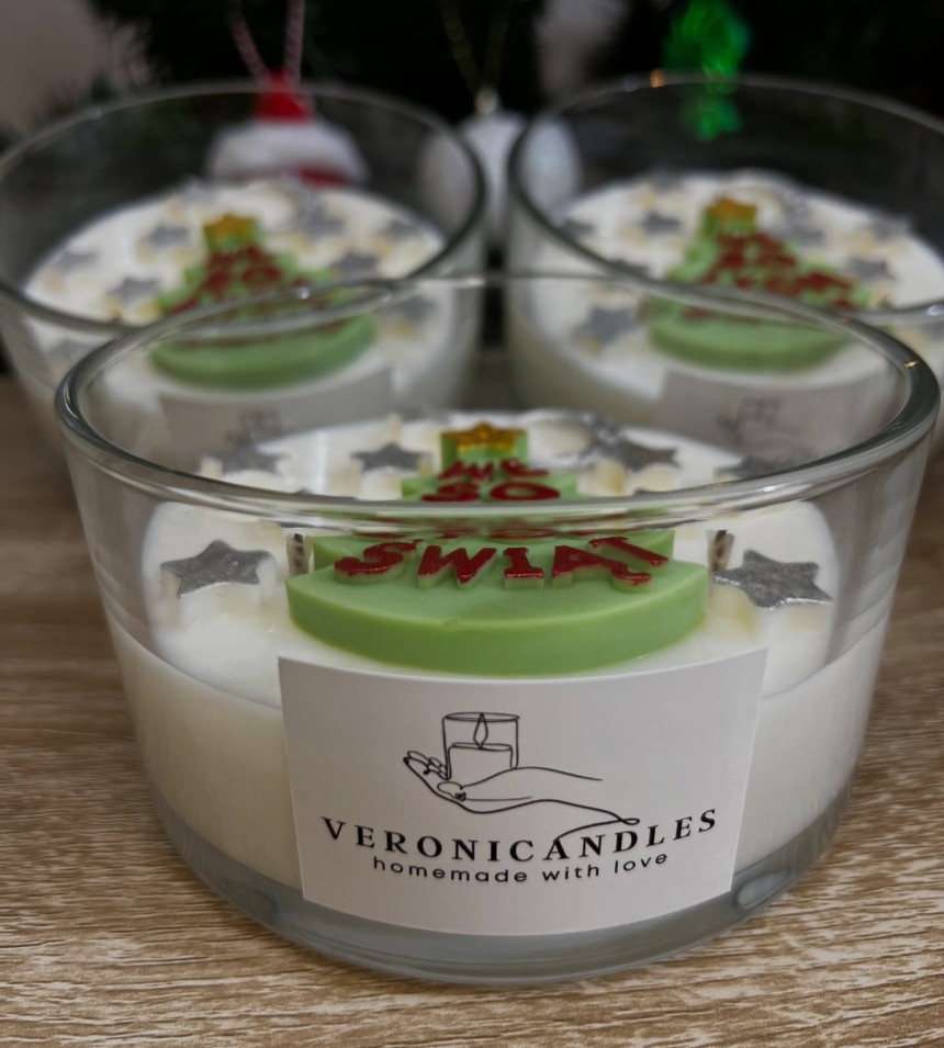 Trzy ręcznie robione świece w szklanych naczyniach z etykietą „Veronicandles – handmade with love”, ozdobione zielonym elementem z napisem „Merry Christmas”, ustawione na drewnianym blacie.