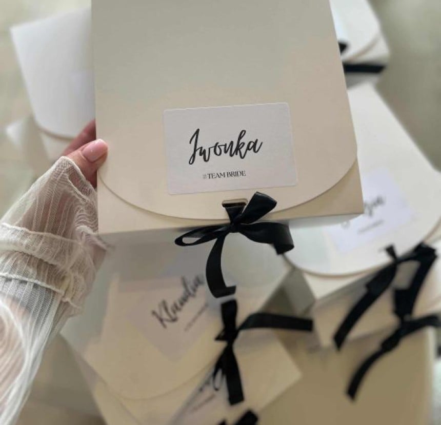 Dłoń trzymająca personalizowane pudełko prezentowe z napisem "Iwonka #TEAM BRIDE", przewiązane czarną wstążką. W tle widać więcej podobnych pudełek.