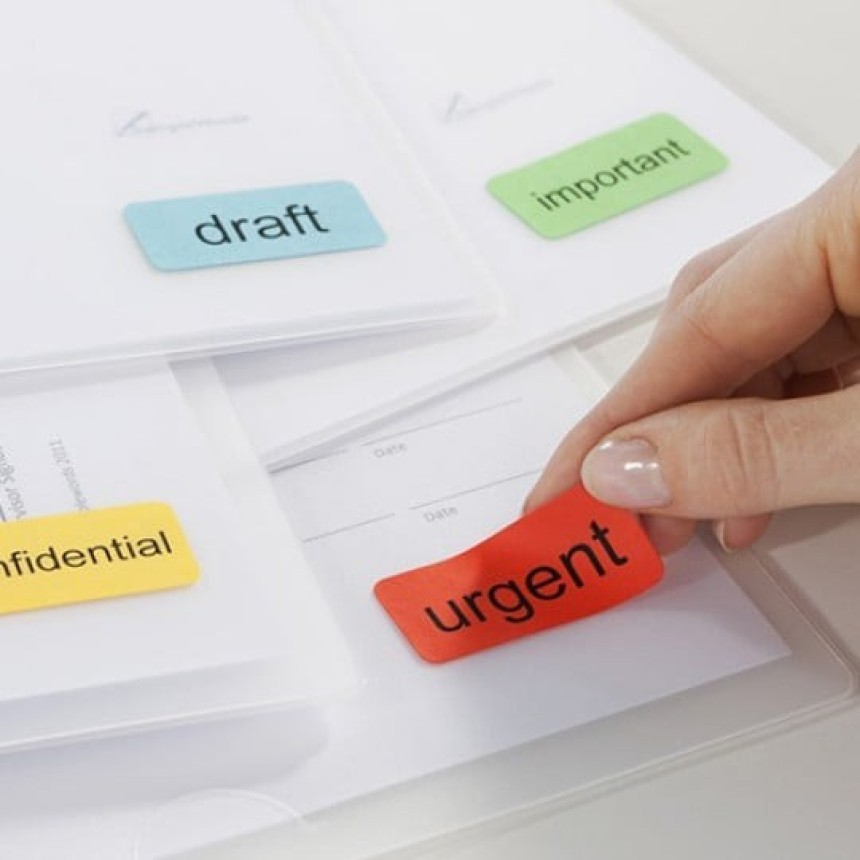 Dłoń przyklejająca czerwoną etykietę z napisem "urgent" na przezroczystą koszulkę na dokumenty. W tle widać inne kolorowe etykiety: "confidential", "draft" i "important".