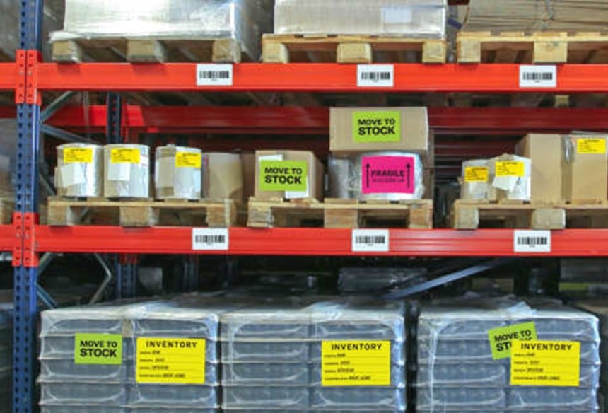 Czerwone regały magazynowe wypełnione paczkami i paletami. Na opakowaniach naklejone są dobrze widoczne, kolorowe etykiety logistyczne z napisami "Move to Stock", "Inventory" oraz "Fragile".