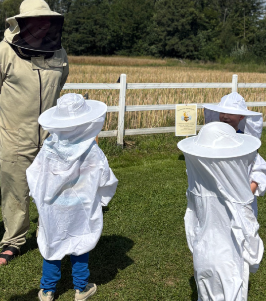 Pszczelarz w beżowym kombinezonie ochronnym stoi na trawie obok dwojga dzieci ubranych w białe kurtki i kapelusze pszczelarskie. W tle widać biały płot, pole uprawne i linię drzew w słoneczny dzień.