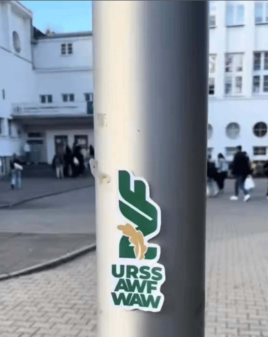 Naklejka z logo URSS AWF WAW przyklejona na słupie przed budynkiem uczelni."