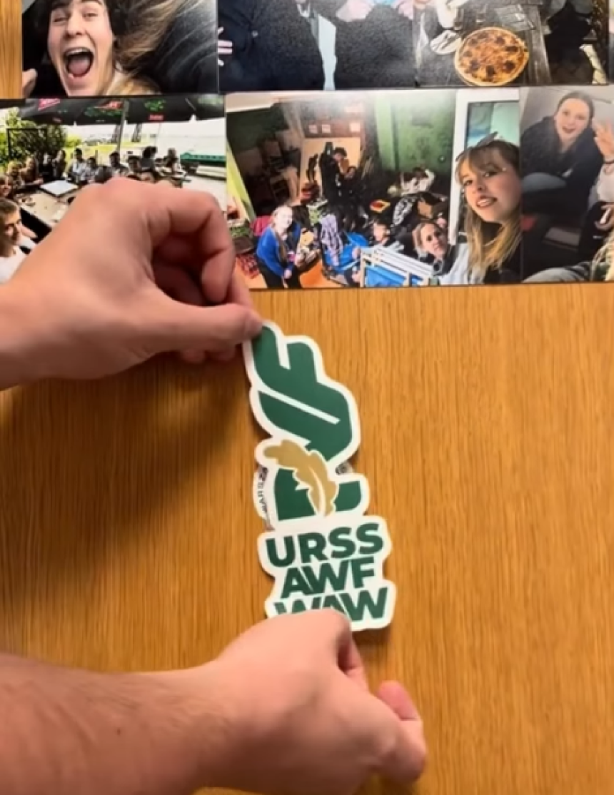 Dłonie przyklejające naklejkę z logo URSS AWF WAW do drewnianej powierzchni obok kolażu ze zdjęć.