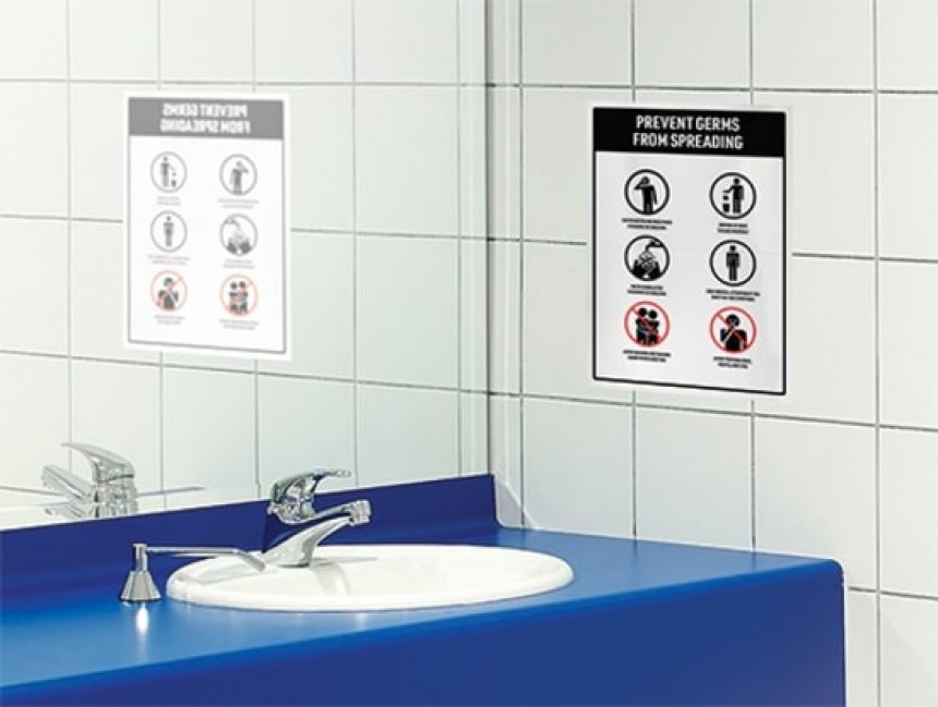 Znak informacyjny z napisem "Prevent Germs From Spreading" i piktogramami dotyczącymi higieny, przyklejony do białej, kafelkowej ściany w łazience.