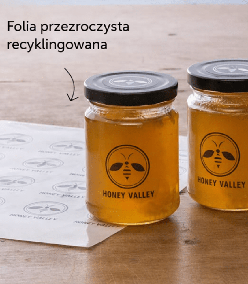 Dwa słoiki miodu z naklejonymi przezroczystymi etykietami "HONEY VALLEY". Obok leży arkusz z etykietami, na który wskazuje strzałka z napisem: "Folia przezroczysta recyklingowana".