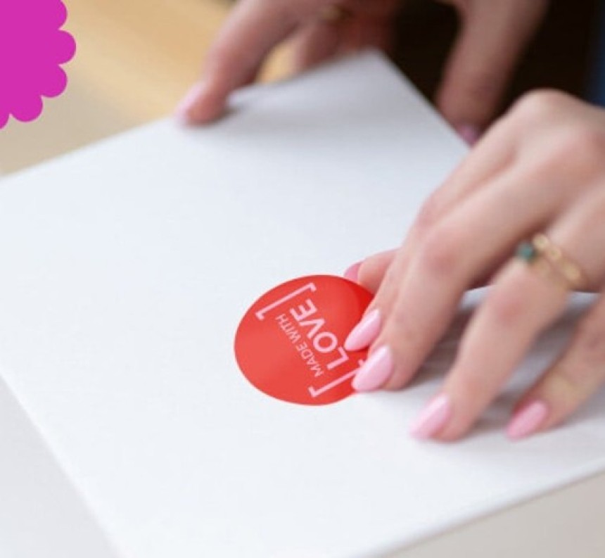 Zbliżenie na dłonie przyklejające na białe pudełko okrągłą, czerwoną naklejkę z napisem „MADE WITH LOVE”.