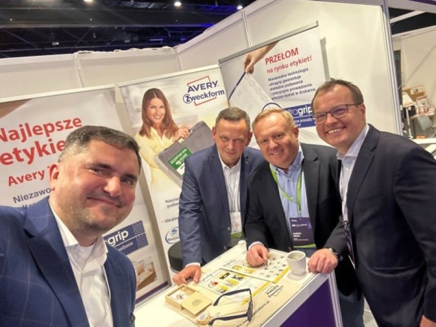 Czterech uśmiechniętych mężczyzn w strojach biznesowych pozuje do zdjęcia typu selfie przy stoisku targowym Avery Zweckform.