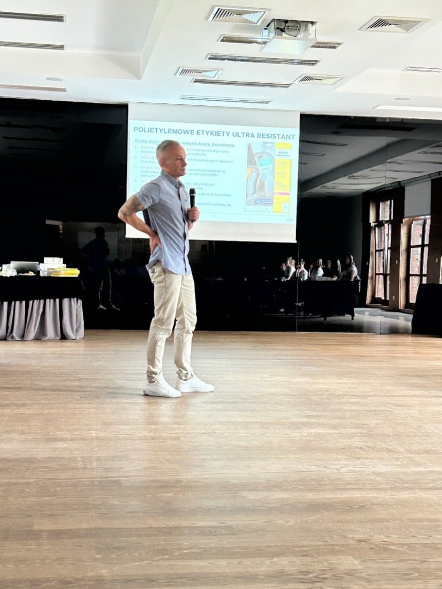 Prelegent w średnim wieku, ubrany w błękitną koszulę i beżowe spodnie, stoi na środku sceny z drewnianą podłogą. W lewej ręce trzyma mikrofon, a prawą ma opartą na biodrze. W tle widać duży ekran projekcyjny ze slajdem o etykietach polietylenowych.