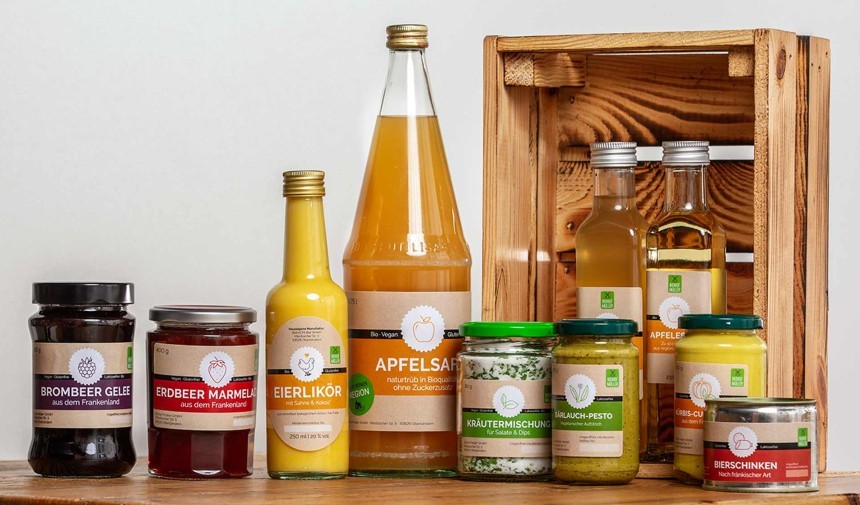 Asortyment przetworów spożywczych marki Biohof Müller. Na drewnianym stole stoją słoiki z dżemem jeżynowym i truskawkowym, pesto, mieszanką ziół, a także butelki z likierem jajecznym, sokiem jabłkowym i olejem. W tle widać drewnianą skrzynkę.