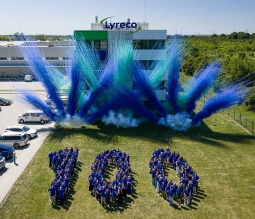 Zespół Lyreco świętuje 100-lecie marki, tworząc na trawie liczbę 100 przed budynkiem firmy. W tle widoczny jest niebiesko-zielony pokaz dymny oraz logo Lyreco na fasadzie budynku.