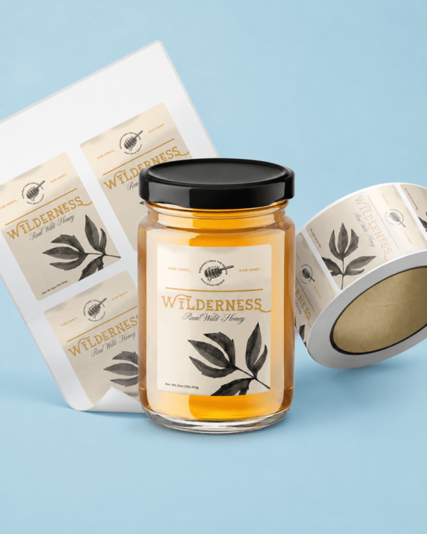 Szklany słoik miodu z etykietą "Wilderness Raw Wild Honey" na jasnoniebieskim tle. Obok leży rolka oraz arkusz z tymi samymi etykietami.