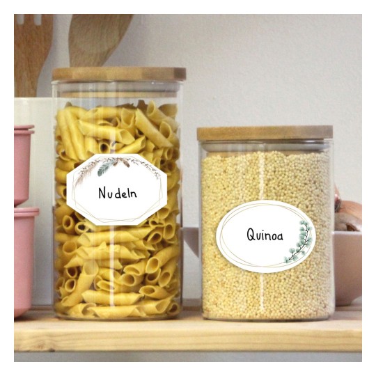 Obraz przedstawia dwa szklane pojemniki na drewnianej półce. Lewy pojemnik zawiera makaron oznaczony jako „Nudeln”, a prawy zawiera komosę ryżową oznaczoną jako „Quinoa”. Oba mają drewniane pokrywki.