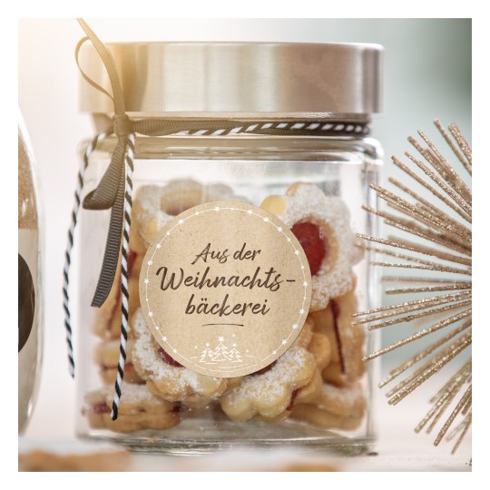 Obrazek przedstawia szklany słoik wypełniony świątecznymi ciasteczkami, z etykietą w języku niemieckim 'Aus der Weihnachtsbäckerei', wskazującą, że pochodzą z bożonarodzeniowej piekarni. Słoik jest elegancko ozdobiony wstążką, a w pobliżu znajdują się dekoracyjne elementy, w tym ozdoba w kształcie gwiazdy.