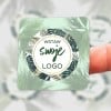 Kwadratowa zielona naklejka z motywem roślinnym i okrągłym kształtem w środku z napisem Wstaw Swoje Logo płożona na palcu