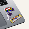 Srebrny laptop z trzema naklejkami: kolorowym napisem ‘FUN’, rysunkiem astronauty w skafandrze oraz żółtą klawiaturą muzyczną z głośnikami