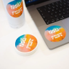 Laptop, butelka i stos naklejek z kolorową grafiką i białym napisem ‘HOLI FEST’ inspirowanym festiwalem kolorów.