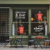 Wejście do kawiarni z dużymi oknami, na których widnieją napisy: ‚KEEP CALM & DRINK COFFEE’ oraz ‚NEED ALWAYS MORE’ wraz z graficznymi kubkami kawy. Przed lokalem stoją stoliki i krzesła w kolorze zielonym i czerwonym