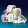 Zdjęcie różnych opakowań suplementów diety i rolek etykiet. Widoczne są m.in. duże plastikowe pojemniki z etykietami ‘Whey Protein’ i ‘Grassfed’, a także rolki naklejek z napisami takimi jak ‘Amino Energy’. Produkty ustawione są na błyszczącej powierzchni z niebieskim gradientowym tłem