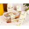 Kwadratowy plastikowy pojemnik z sałatką, oznaczony zielono-pomarańczową etykietą „Organic Burrito Bowl”, leży na jasnym blacie; w tle widoczne rolki z identycznymi etykietami i owoce.