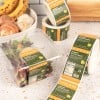 Kwadratowy plastikowy pojemnik z sałatką, oznaczony zielono-pomarańczową etykietą „Organic Burrito Bowl”, leży na jasnym blacie; w tle widoczne rolki z identycznymi etykietami i owoce.