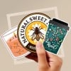 Dłoń trzyma trzy kolorowe naklejki: pierwsza przedstawia kubek z napojem, druga okrągła z rysunkiem pszczoły i napisem 'Natural Sweet', trzecia w kształcie filiżanki kawy z motywem roślinnym i napisem 'Roast coffee house