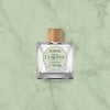 Kwadratowy flakon z drewnianą nakrętką. Etykieta produktu nosi nazwę „Beard Oil Petite Coquette Lavender Dream” na jasnozielonym tle. Flakon ustawiony na blacie o wzorze jasnego marmuru.