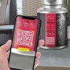 Dłoń trzymająca smartfon z włączonym aparatem skanującym duży czerwony kod QR na etykiecie suplementu diety, w tle widoczna butelka z tą samą etykietą, co pokazuje użycie kodu do uzyskania informacji o multiwitaminach