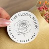 Okrągła naklejka z logo "The Floral Nook" trzymana między palcami. Na białym tle widnieje czarna grafika liniowa róży w centrum, otoczona tekstem "THE FLORAL NOOK" u góry i "FLORISTRY • SINCE 2021" u dołu. W tle widoczne suszone kwiaty.