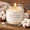 Zapalona świeca zapachowa w przezroczystym, szklanym słoiku, stojąca na rustykalnej, drewnianej powierzchni. Na białej etykiecie widnieje napis 'Cotton & Lavender, Natural Soy Candle, Hand Poured'. Wokół słoika i w tle ułożone są puszyste kwiaty bawełny, tworząc ciepłą, przytulną atmosferę.