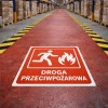 Duża, czerwona naklejka podłogowa z napisem "DROGA PRZECIWPOŻAROWA" i piktogramem ewakuacji, umieszczona na wyznaczonej drodze w korytarzu magazynowym.