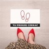 Widok z góry na stopy w czerwonych szpilkach stojące przed białą naklejką podłogową. Na naklejce znajduje się napis "TU PROSZĘ CZEKAĆ" na czarnym pasku oraz grafika z czerwonym obrysem stóp.