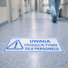 Podłużna naklejka podłogowa na korytarzu szpitalnym. Na naklejce znajduje się niebieski piktogram ostrzegawczy (trójkąt z wykrzyknikiem) i napis: "UWAGA PRZEJŚCIE TYLKO DLA PERSONELU". W tle widać idącą postać w stroju medycznym.
