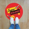 Stopy osoby w białych trampkach na tle dużej, czerwonej, okrągłej naklejki podłogowej. Na naklejce widnieje duży, stylizowany napis "Super SOLDES" (Super Wyprzedaż). Naklejka przyklejona jest do drewnianej podłogi.