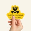 Dłoń trzymająca żółtą naklejkę o niestandardowym kształcie, z czarnym logo i napisem "Custom Creations Design & Print".