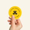 Dłoń trzymająca pojedynczą, okrągłą, żółtą naklejkę z czarnym logo i napisem "Custom Creations Design & Print".