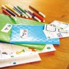 Obraz przedstawia kolorowe zeszyty i markery oraz edukacyjne karty. Układ obejmuje zielone i niebieskie zeszyty oraz różnorodne kolorowe długopisy na drewnianej powierzchni.