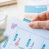 Obraz przedstawia rękę trzymającą stronę z planera z miesiącem październikiem. Strona ma naklejkę kwiatową, etykietę z napisem 'OFFLINE' oraz różne oznaczone sekcje dla dni tygodnia, wskazujące zaplanowane aktywności.