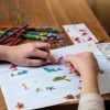 Obraz przedstawia dwie ręce pracujące nad kolorową kolażem o tematyce oceanicznej. Jedna ręka nakleja naklejkę syrenki, podczas gdy na papierze są ułożone różne naklejki zwierząt wodnych, w tym delfinów i żółwi. Wokół miejsca pracy znajdują się kolorowe kredki i dodatkowe naklejki, co tworzy zabawną i kreatywną atmosferę.