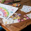 Obraz przedstawia kreatywną przestrzeń roboczą z kolorowymi rysunkami i różnymi naklejkami. Na papierze narysowana jest tęcza, ozdobiona błyszczącymi klejnotami. Na drewnianym stole znajduje się kilka arkuszy naklejek z postaciami z kreskówek, sercami i kwiatami, a także kolorowe kredki i cukierki.